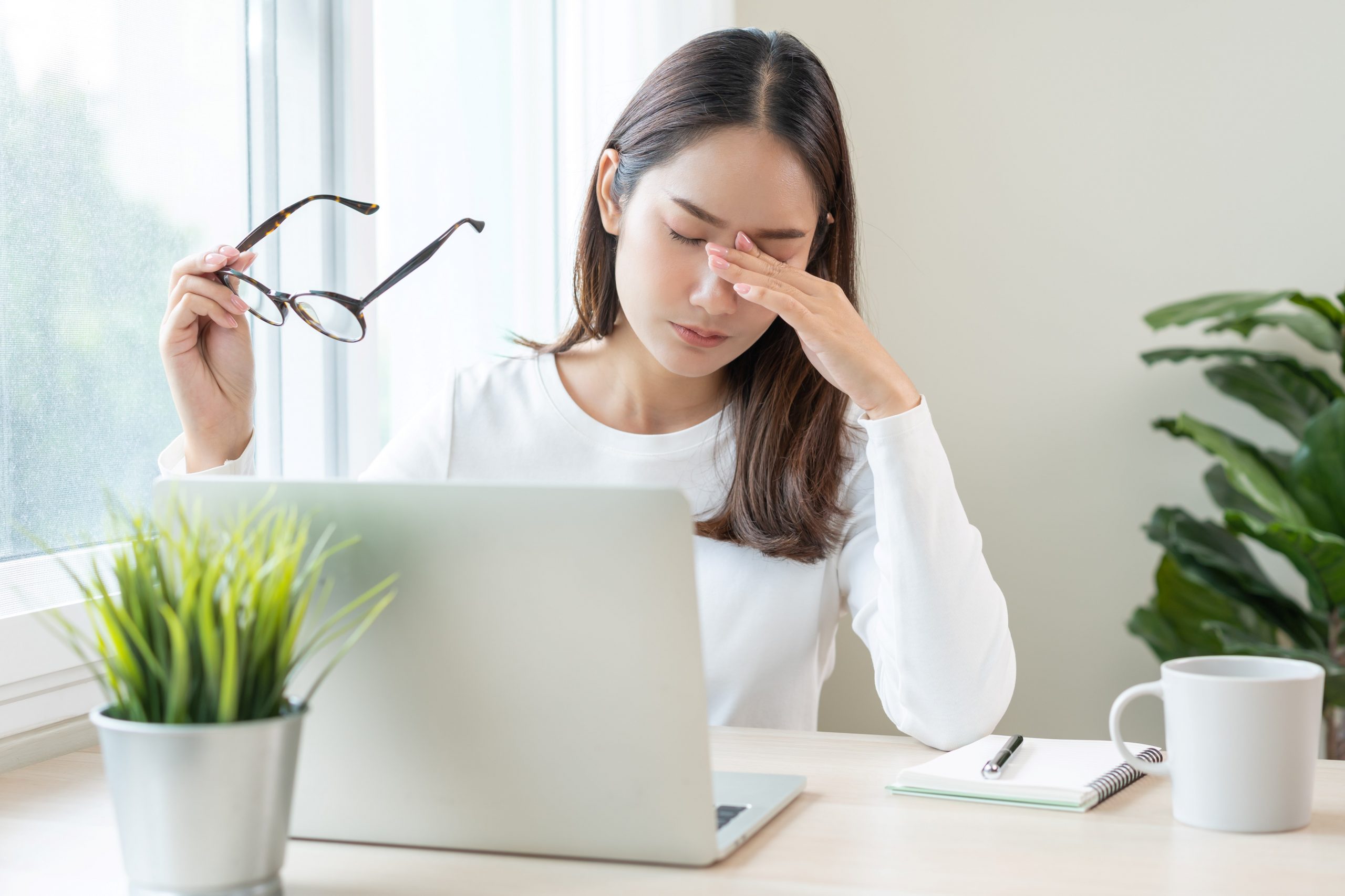 Eine Frau sitzt am Schreibtisch mit Laptop, hält ihre Brille in der Hand und reibt sich die Augen. Auf dem Tisch stehen eine Tasse, ein Notizbuch und eine Pflanze. Das Bild symbolisiert Augenbelastung oder Schwierigkeiten mit der Gleitsichtbrille.