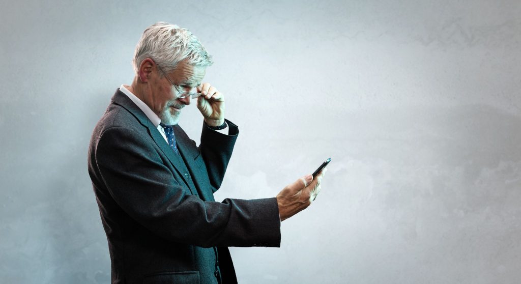 Älterer Mann hält sein Smartphone auf Abstand und setzt eine Lesebrille auf – typisches Zeichen für Altersweitsichtigkeit (Presbyopie) im Alltag